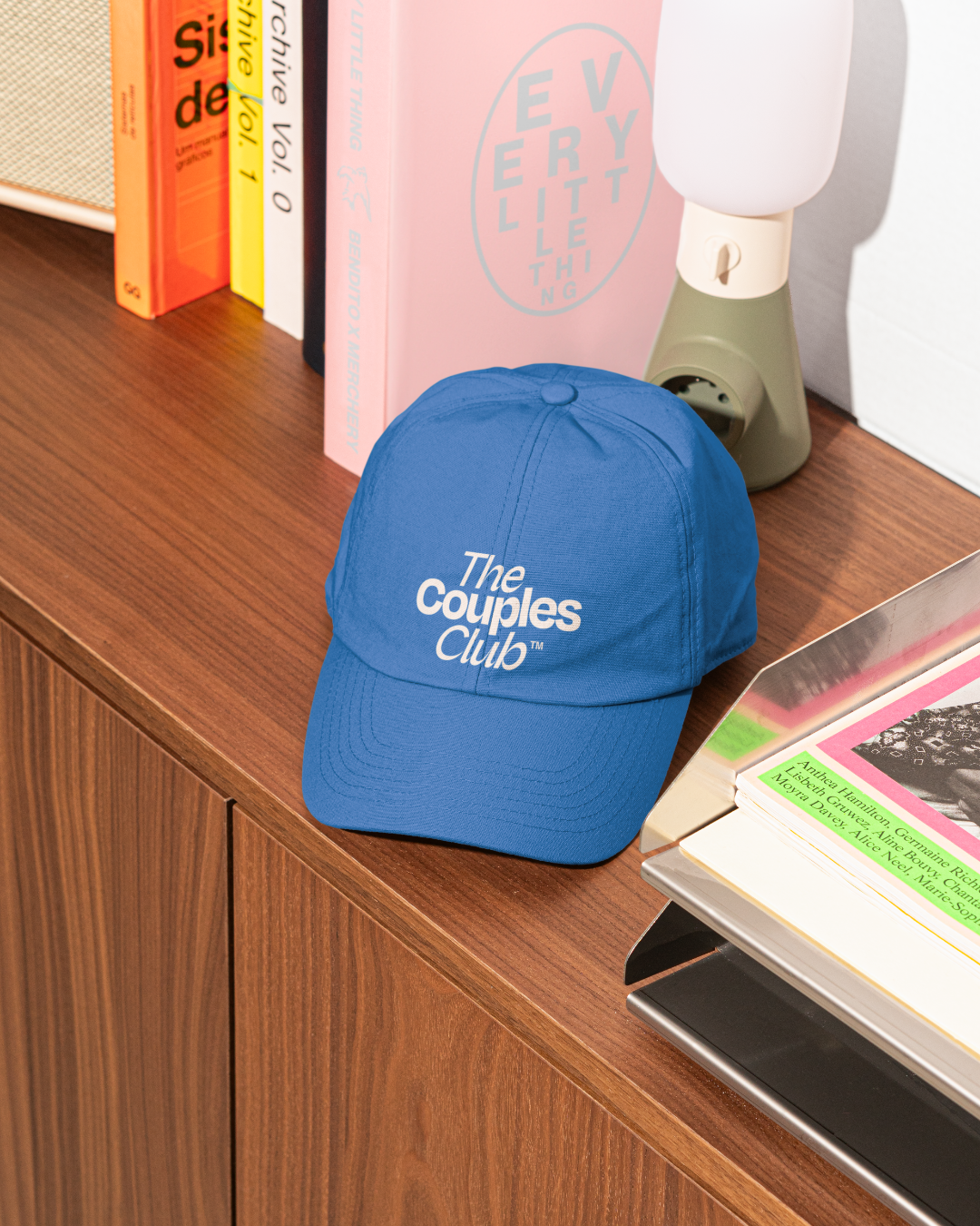 Blue cap with 'The Couples Club' text on a wooden surface with books and a lamp in the background