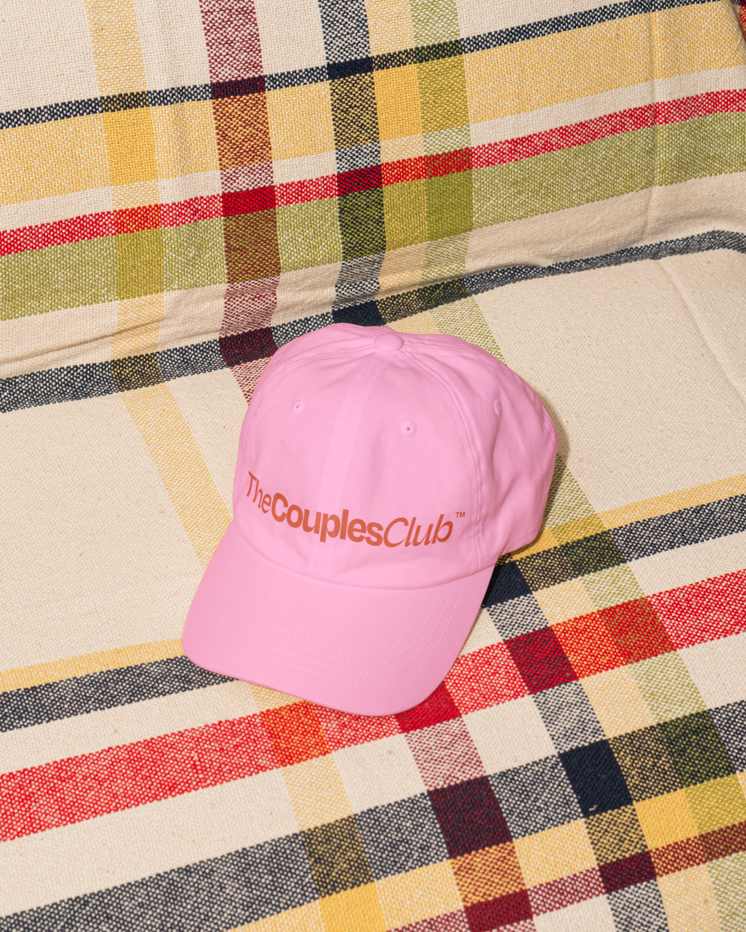Pink cap with 'The Couples Club' logo on a plaid fabric background