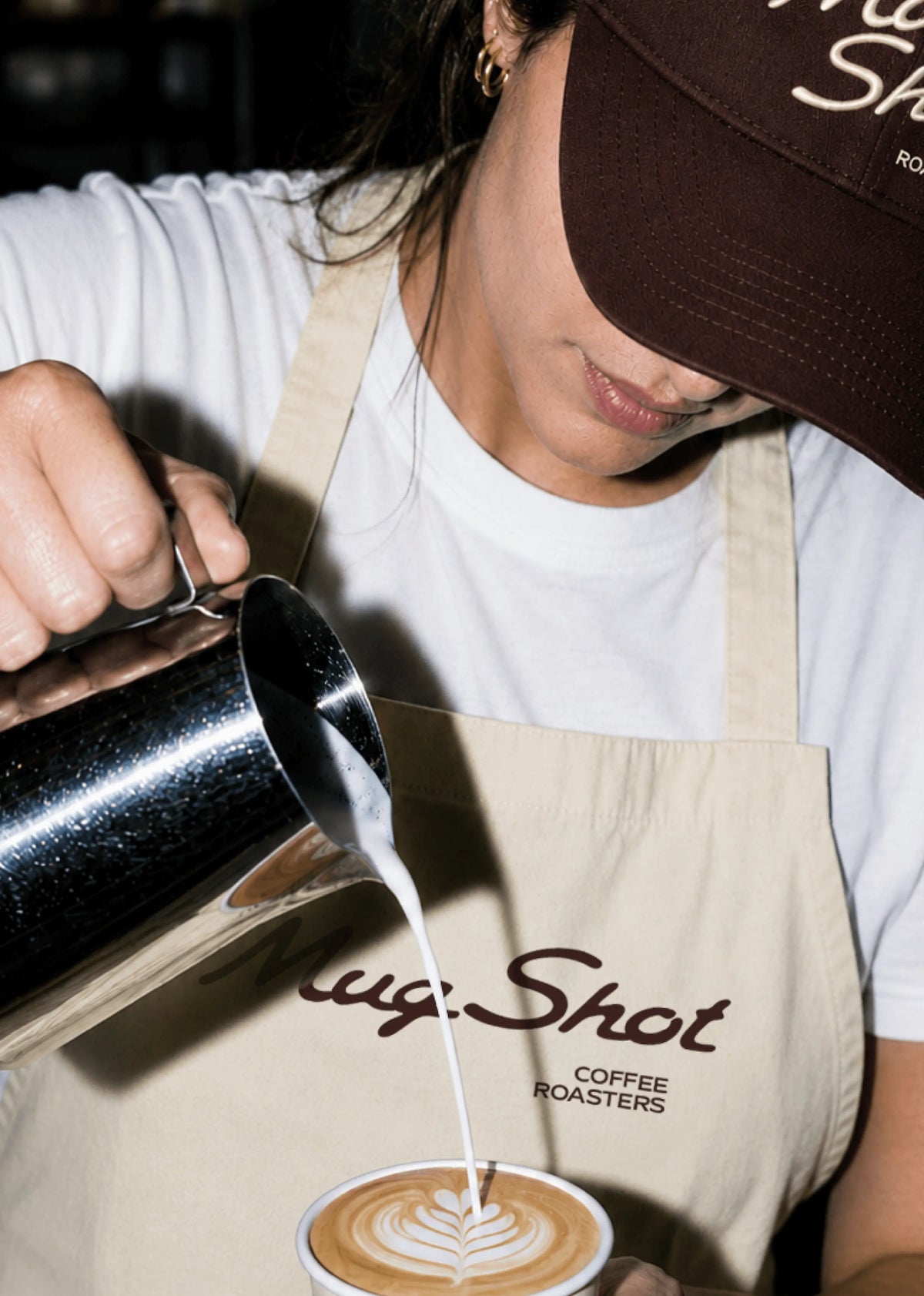 Barista in a Mug Shot branded apron and cap pouring latte art into a cup