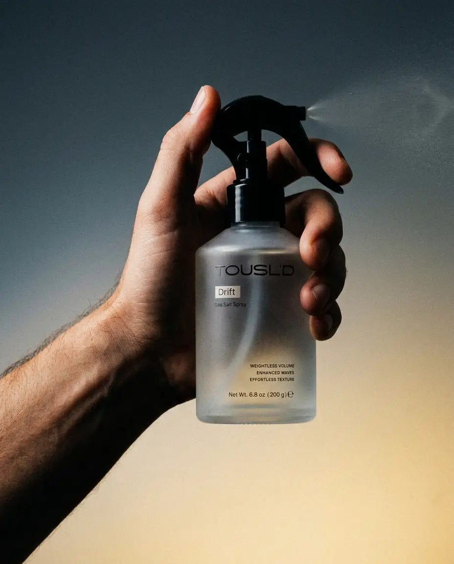 Hand holding a Tousl’d spray bottle against a gradient background